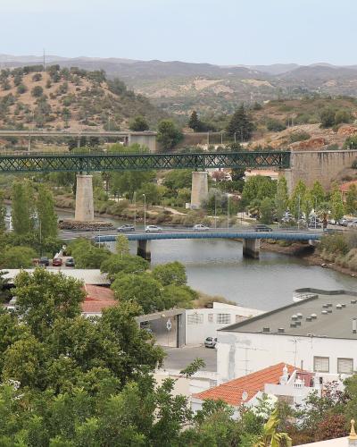 A high-angle view of a river valley in a Portuguese town featuring three distinct bridges: a stone aqueduct with a green metal truss section, a lower concrete road bridge, and a modern highway viaduct in the background.