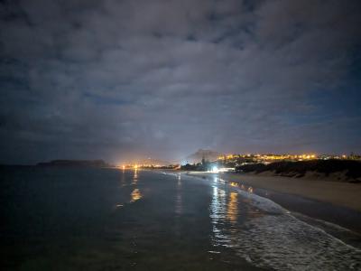 A night-time photograph of a wide sandy beach with gentle waves, showing distant coastal town lights and a prominent mountain silhouette in the background under a cloudy sky.