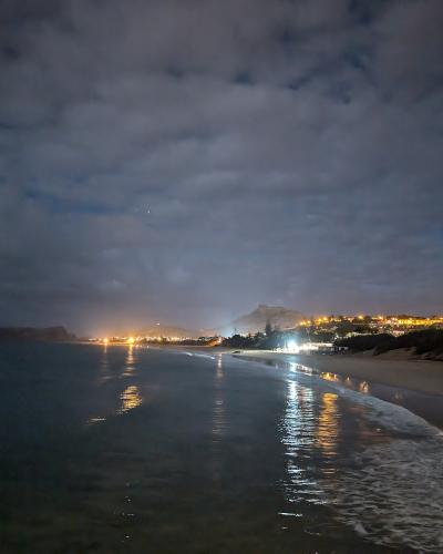 A night-time photograph of a wide sandy beach with gentle waves, showing distant coastal town lights and a prominent mountain silhouette in the background under a cloudy sky.