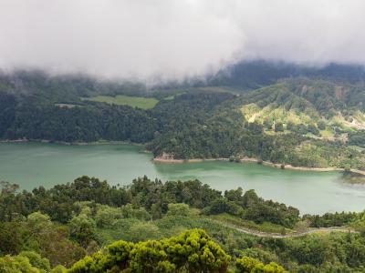 A panoramic landscape view of a large, green-tinted freshwater lake surrounded by lush, forested volcanic hills and a dam structure on the right side.