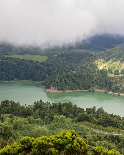A panoramic landscape view of a large, green-tinted freshwater lake surrounded by lush, forested volcanic hills and a dam structure on the right side.