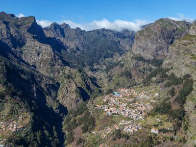 A high-angle landscape photograph showing a village with terracotta-roofed houses nestled in a deep, steep-sided mountain valley surrounded by rugged peaks.