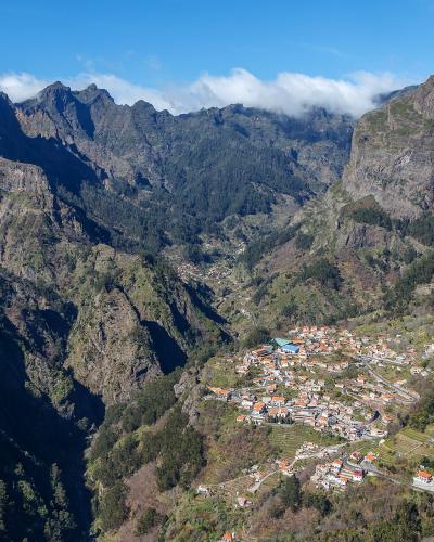 A high-angle landscape photograph showing a village with terracotta-roofed houses nestled in a deep, steep-sided mountain valley surrounded by rugged peaks.