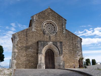 A low-angle photograph of a medieval stone church facade featuring a large rose window, a pointed arched doorway with carved columns, and a gabled roof against a blue sky.