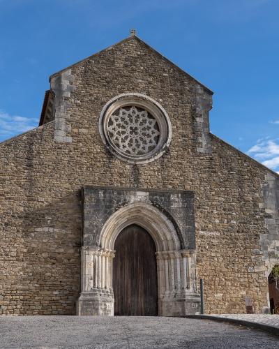 A low-angle photograph of a medieval stone church facade featuring a large rose window, a pointed arched doorway with carved columns, and a gabled roof against a blue sky.