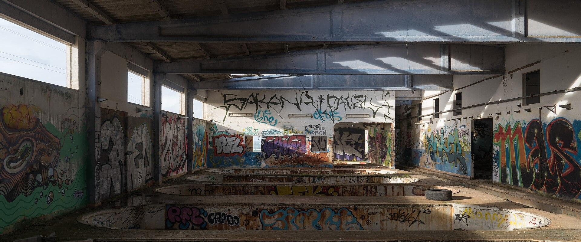 An interior view of a derelict industrial building featuring large, circular concrete tanks or basins, with walls and surfaces heavily covered in colorful graffiti and skylights in the roof.