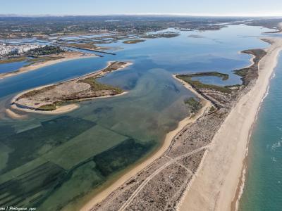 An aerial photograph showing a long, narrow sandy barrier island separating a lagoon system from the ocean, with a town visible in the background across the water.