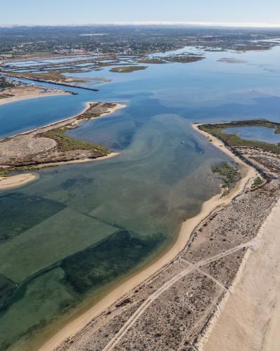 An aerial photograph showing a long, narrow sandy barrier island separating a lagoon system from the ocean, with a town visible in the background across the water.