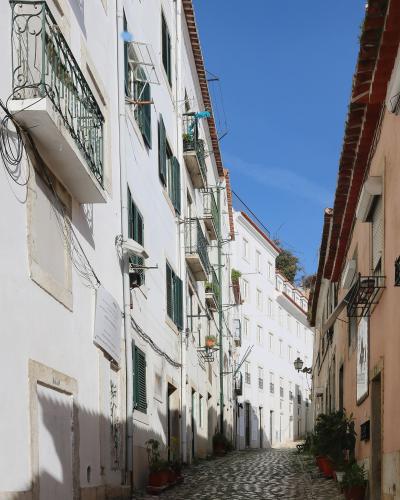 A narrow, upward-sloping cobblestone street lined with white-washed buildings featuring green shutters and wrought-iron balconies under a clear blue sky.