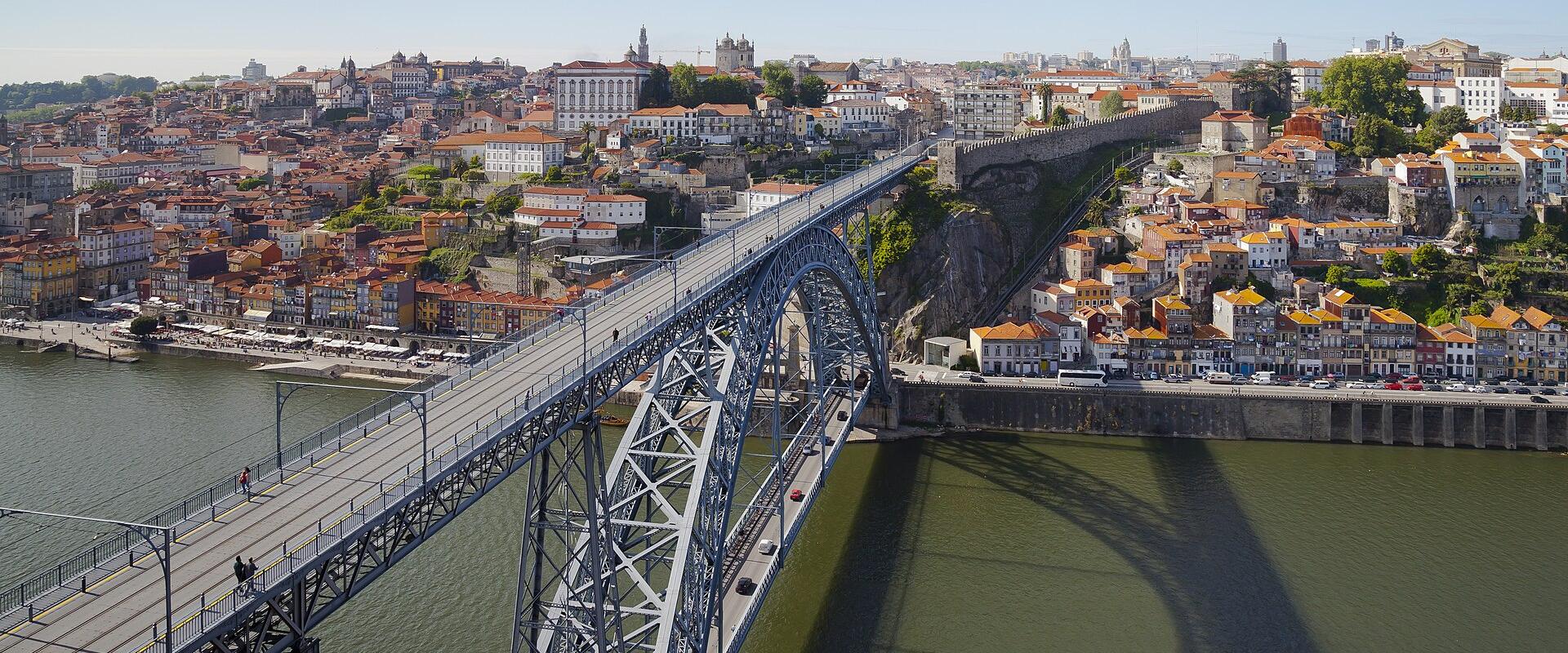 A high-angle view of a large, double-deck metal arch bridge spanning a wide river, with a cityscape of terracotta-roofed buildings on the far bank and a stone embankment on the near bank.