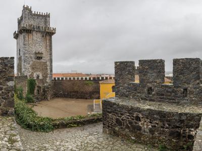 A stone courtyard within a medieval fortress featuring a prominent square keep with crenellations, stone ramparts, and two flags flying on the right side.