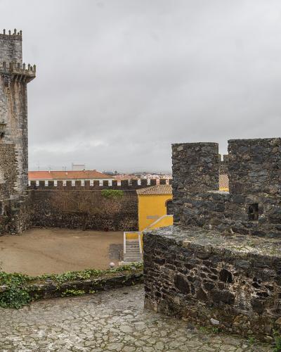 A stone courtyard within a medieval fortress featuring a prominent square keep with crenellations, stone ramparts, and two flags flying on the right side.
