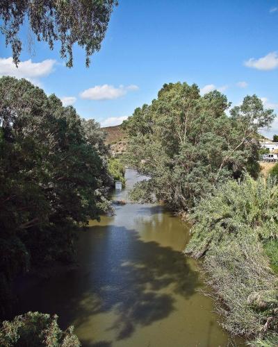 A calm river flowing through a lush, tree-lined valley under a blue sky, with a watermark in the bottom right corner reading "Portuguese_eyes / Vitor Oliveira".