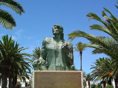 A bronze statue of a standing man on a stone pedestal with a plaque reading "CRISTOVÃO COLOMBO", surrounded by palm trees and white buildings under a blue sky.