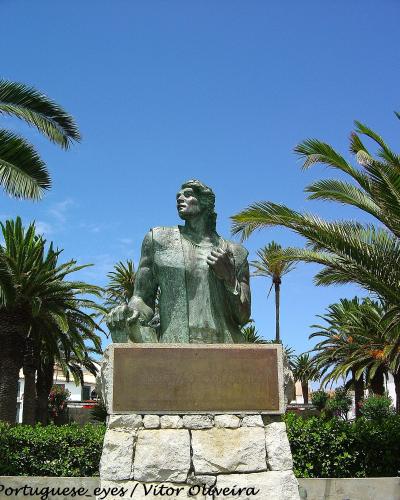 A bronze statue of a standing man on a stone pedestal with a plaque reading "CRISTOVÃO COLOMBO", surrounded by palm trees and white buildings under a blue sky.