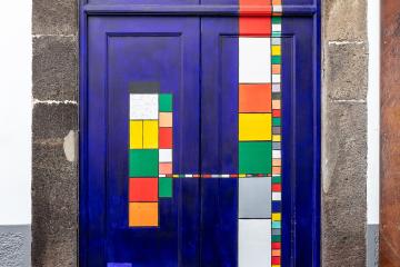 A close-up of a vibrant blue double door with colorful geometric patterns, framed by dark stone, featuring a small plaque reading "S. CASA DA N.º 39" and a sign for "MUNDIAL SAPATARIAS" at the bottom.
