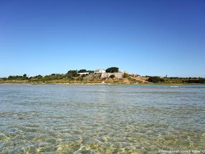 A view across shallow, clear water towards a coastal hilltop settlement featuring white buildings and a stone fortification under a clear blue sky.