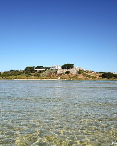A view across shallow, clear water towards a coastal hilltop settlement featuring white buildings and a stone fortification under a clear blue sky.