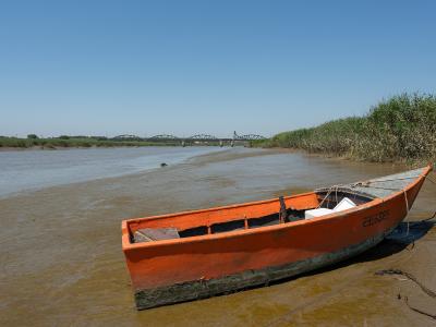 An orange wooden boat rests on a muddy riverbank in the foreground, with a large green truss bridge spanning the water in the distance under a clear blue sky.