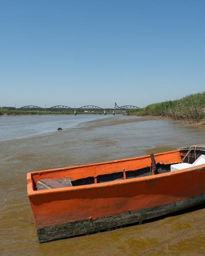An orange wooden boat rests on a muddy riverbank in the foreground, with a large green truss bridge spanning the water in the distance under a clear blue sky.