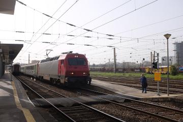 A red and silver electric locomotive, number 5618-1, pulling passenger carriages along a multi-track railway line with overhead catenary wires, under an overcast sky at a station platform.