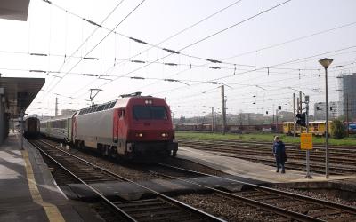 A red and silver electric locomotive, number 5618-1, pulling passenger carriages along a multi-track railway line with overhead catenary wires, under an overcast sky at a station platform.