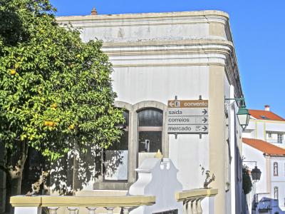 A white building with yellow trim on a cobblestone street corner, featuring a directional signpost pointing to 'convento', 'saida', 'correios', and 'mercado', and a small plaque on the wall reading 'galeria sto antónio'.