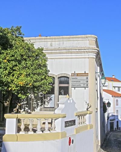 A white building with yellow trim on a cobblestone street corner, featuring a directional signpost pointing to 'convento', 'saida', 'correios', and 'mercado', and a small plaque on the wall reading 'galeria sto antónio'.