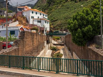 A concrete channelized stream (ribeira) running through a village, flanked by stone retaining walls, with a green metal railing in the foreground and white houses with terracotta roofs on a hillside in the background.
