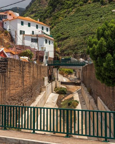 A concrete channelized stream (ribeira) running through a village, flanked by stone retaining walls, with a green metal railing in the foreground and white houses with terracotta roofs on a hillside in the background.