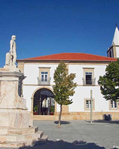 A marble statue of a standing figure on a tall pedestal in a paved town square, with a white building featuring a red-tiled roof and a bell tower in the background.