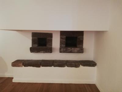 An interior shot of a white-washed wall featuring two small, square stone openings (likely ovens or fireplaces) above a long stone bench, set on a wooden floor.