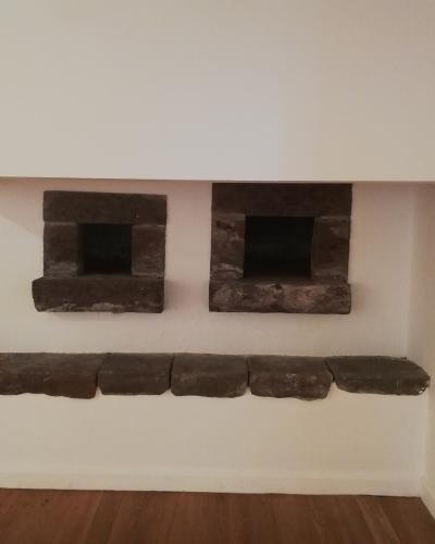 An interior shot of a white-washed wall featuring two small, square stone openings (likely ovens or fireplaces) above a long stone bench, set on a wooden floor.