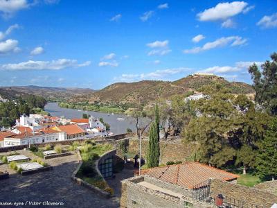 A high-angle view from a stone terrace overlooking the town of Alcoutim, the Guadiana River, and the Alcoutim Castle ruins on a distant hill.