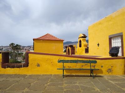 A view of a bright yellow-walled historic fortification with a green bench in the foreground, overlooking a coastal city and hills.