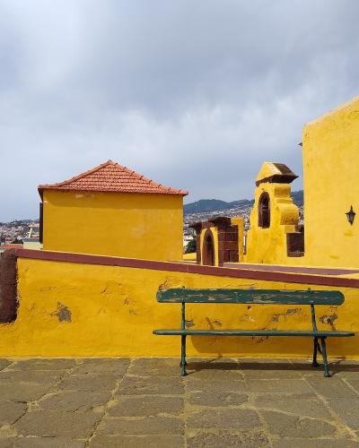 A view of a bright yellow-walled historic fortification with a green bench in the foreground, overlooking a coastal city and hills.