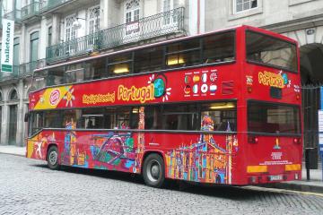 A red double-decker sightseeing bus with "City Sightseeing Portugal" branding and colorful illustrations of Portuguese landmarks, parked on a cobblestone street in front of a building with a "balletteatro" sign.