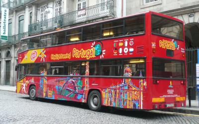 A red double-decker sightseeing bus with "City Sightseeing Portugal" branding and colorful illustrations of Portuguese landmarks, parked on a cobblestone street in front of a building with a "balletteatro" sign.