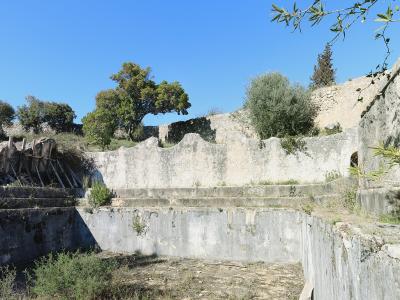 The image shows a weathered, empty concrete basin or reservoir structure with stepped walls, surrounded by trees and stone ruins under a blue sky.