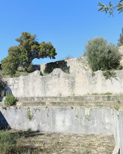 The image shows a weathered, empty concrete basin or reservoir structure with stepped walls, surrounded by trees and stone ruins under a blue sky.