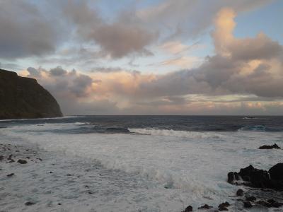 The image shows a rugged, volcanic coastline with dark rocks and steep cliffs meeting the Atlantic Ocean under a cloudy sky at dusk or dawn.