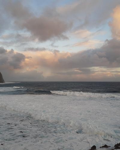 The image shows a rugged, volcanic coastline with dark rocks and steep cliffs meeting the Atlantic Ocean under a cloudy sky at dusk or dawn.