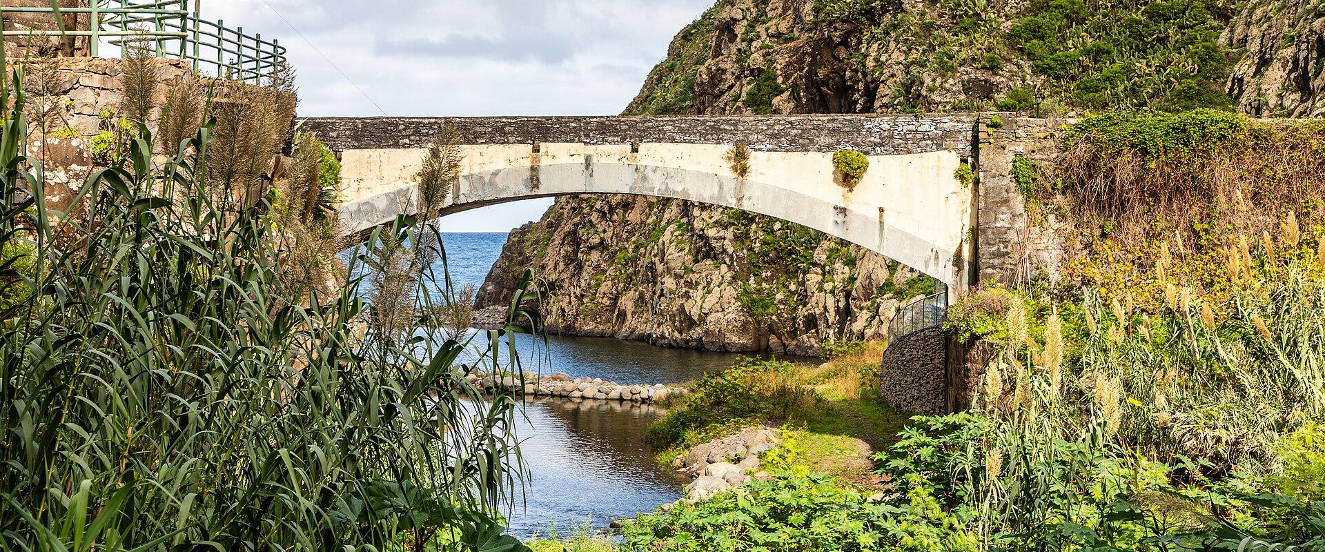 A stone arch bridge spanning a small inlet or river mouth where it meets the ocean, with lush green vegetation and a steep rocky hillside in the background.