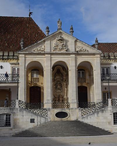 A low-angle photograph of the ornate stone facade of the Chapel of St. John the Baptist (Capela de São João Baptista), featuring three arched doorways, a central sculpted niche, and a coat of arms above the pediment, with a wide staircase leading up 