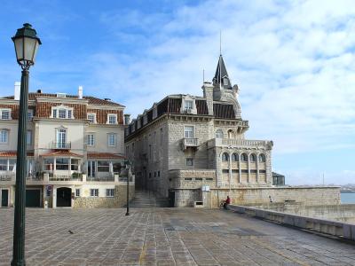 A waterfront promenade in Cascais, Portugal, featuring the stone Palácio dos Condes de Castro Guimarães on the right and the adjacent Palácio de Seixas on the left.