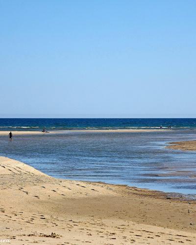 A wide sandy beach with a tidal channel or inlet flowing towards the ocean, featuring a few people walking on the sand and a sailboat in the distance under a clear blue sky.