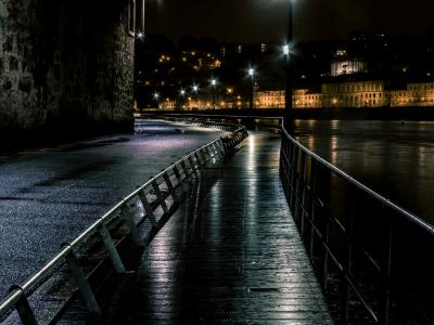 A wet, wooden boardwalk with metal railings at night, running alongside a stone wall on the left and a river on the right, with illuminated historic buildings visible across the water.