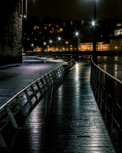 A wet, wooden boardwalk with metal railings at night, running alongside a stone wall on the left and a river on the right, with illuminated historic buildings visible across the water.