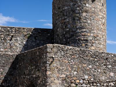 A close-up view of a medieval stone fortress featuring a large cylindrical tower and a lower wall with an arched gateway, set against a blue sky.