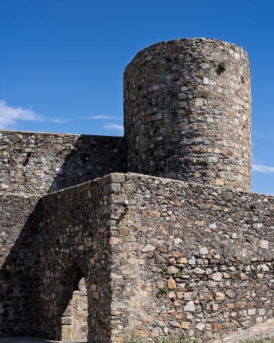 A close-up view of a medieval stone fortress featuring a large cylindrical tower and a lower wall with an arched gateway, set against a blue sky.